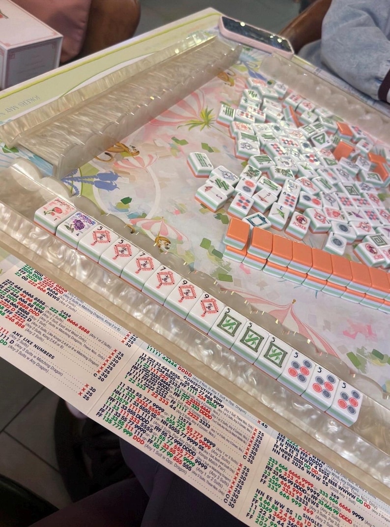 May include: A Mahjong set with white and orange tiles, arranged on a floral patterned surface. The tiles display various symbols and numbers, with a clear plastic tray for the tiles and a printed game guide.