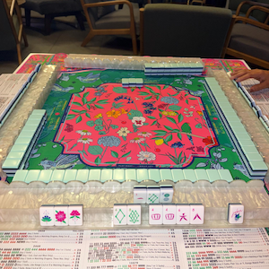 May include: A Mahjong set featuring a vibrant pink floral design on the playing surface. The set includes white tiles with symbols, racks, and a printed score sheet. The game is set up and ready to be played.