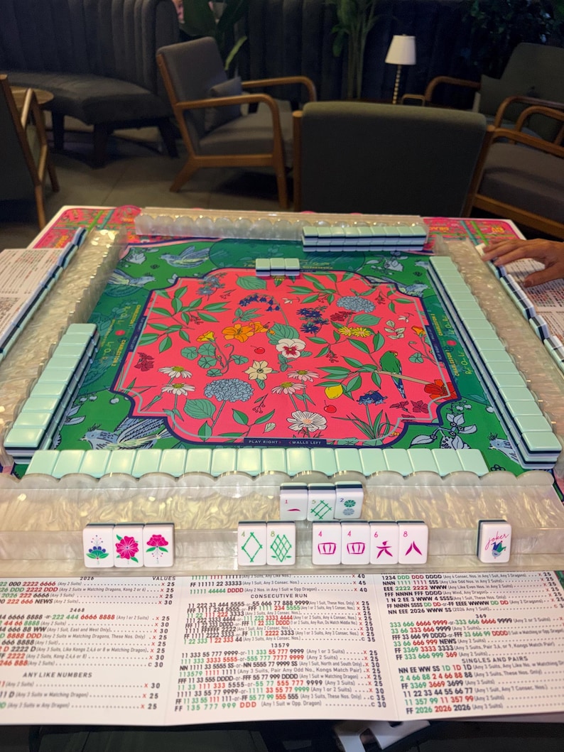 May include: A Mahjong set featuring a vibrant pink floral design on the playing surface. The set includes white tiles with symbols, racks, and a printed score sheet. The game is set up and ready to be played.