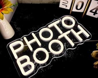 May include: A clear acrylic sign with the words "PHOTO BOOTH" in white, illuminated with a soft glow. The sign has a wavy outline and is set against a dark background. A calendar shows the date July 4th.