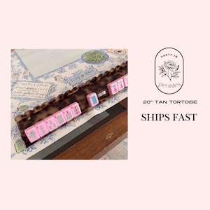May include: A tan tortoise mahjong tile rack with pink tiles. The tiles are arranged on a decorative tray with a scalloped edge. The background features a floral pattern and the text "Party in Peonies".