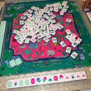 May include: A Mahjong set with white tiles featuring green and red symbols, scattered on a floral patterned mat. The mat has text that says "Charleston". A row of tiles with floral and joker designs are visible.
