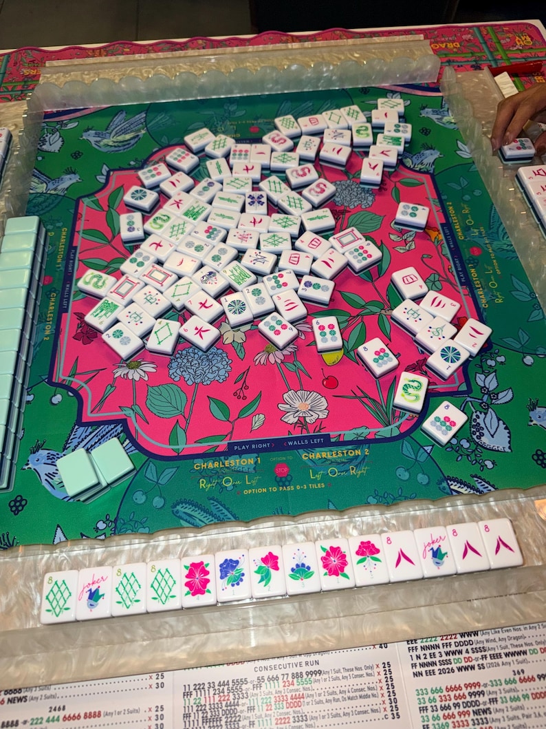 May include: A Mahjong set with white tiles featuring green and red symbols, scattered on a floral patterned mat. The mat has text that says "Charleston". A row of tiles with floral and joker designs are visible.
