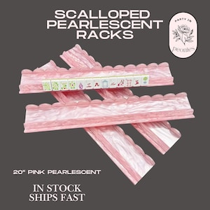 May include: Four 50.8 cm pink pearlescent mahjong racks with a scalloped top edge. The racks are arranged in a crisscross pattern. One rack holds a row of twelve mahjong tiles with various green, red, and blue symbols and characters printed on them.