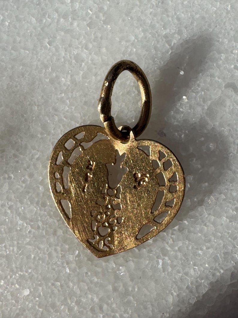 Puede incluir: Un colgante de coraz&oacute;n de oro con un delicado dise&ntilde;o de corte. El colgante presenta dos figuras abraz&aacute;ndose dentro del coraz&oacute;n.