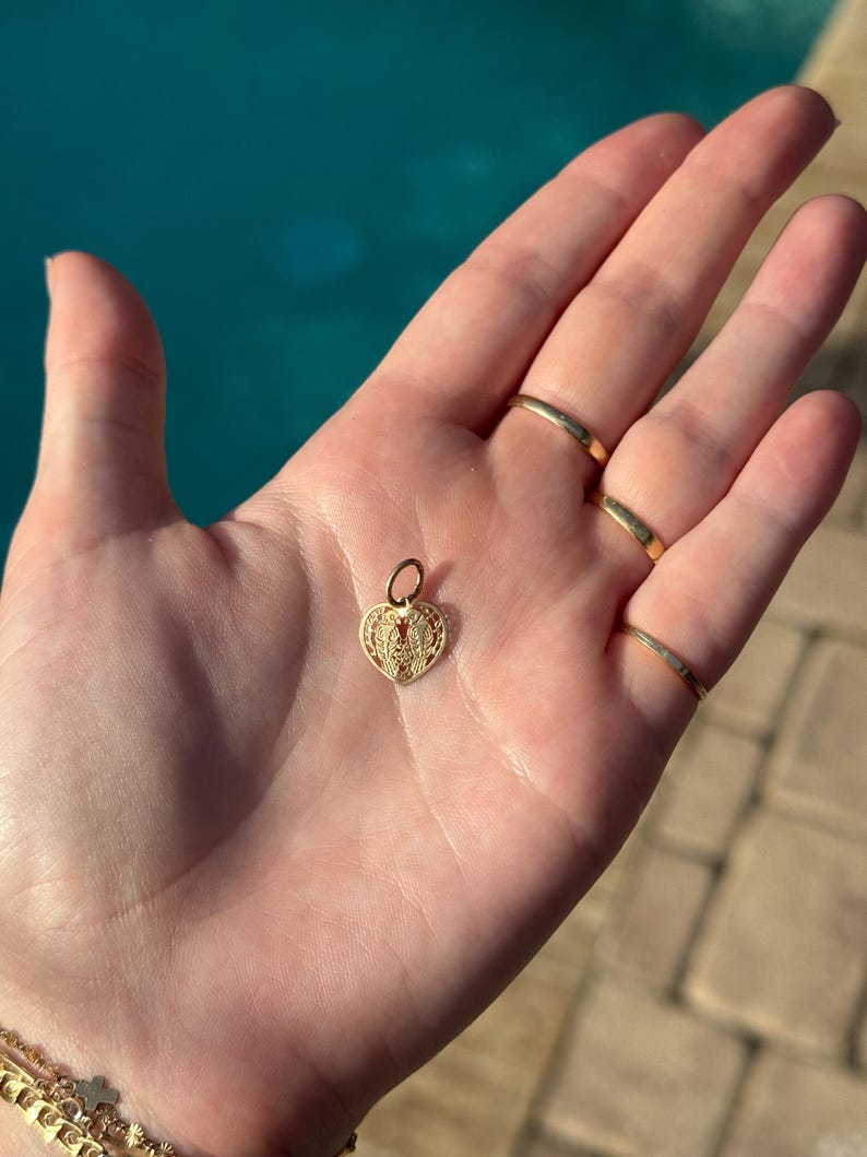 Puede incluir: Un dije de coraz&oacute;n de oro con intrincados detalles de filigrana. El dije cuelga de un peque&ntilde;o lazo de oro.