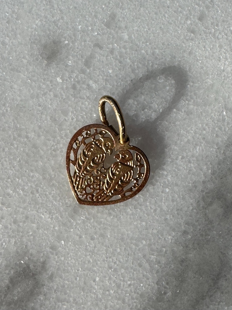 Puede incluir: Un colgante de coraz&oacute;n de oro con un delicado dise&ntilde;o de filigrana que presenta dos p&aacute;jaros entrelazados. El colgante tiene un bucle en la parte superior para sujetarlo a una cadena.