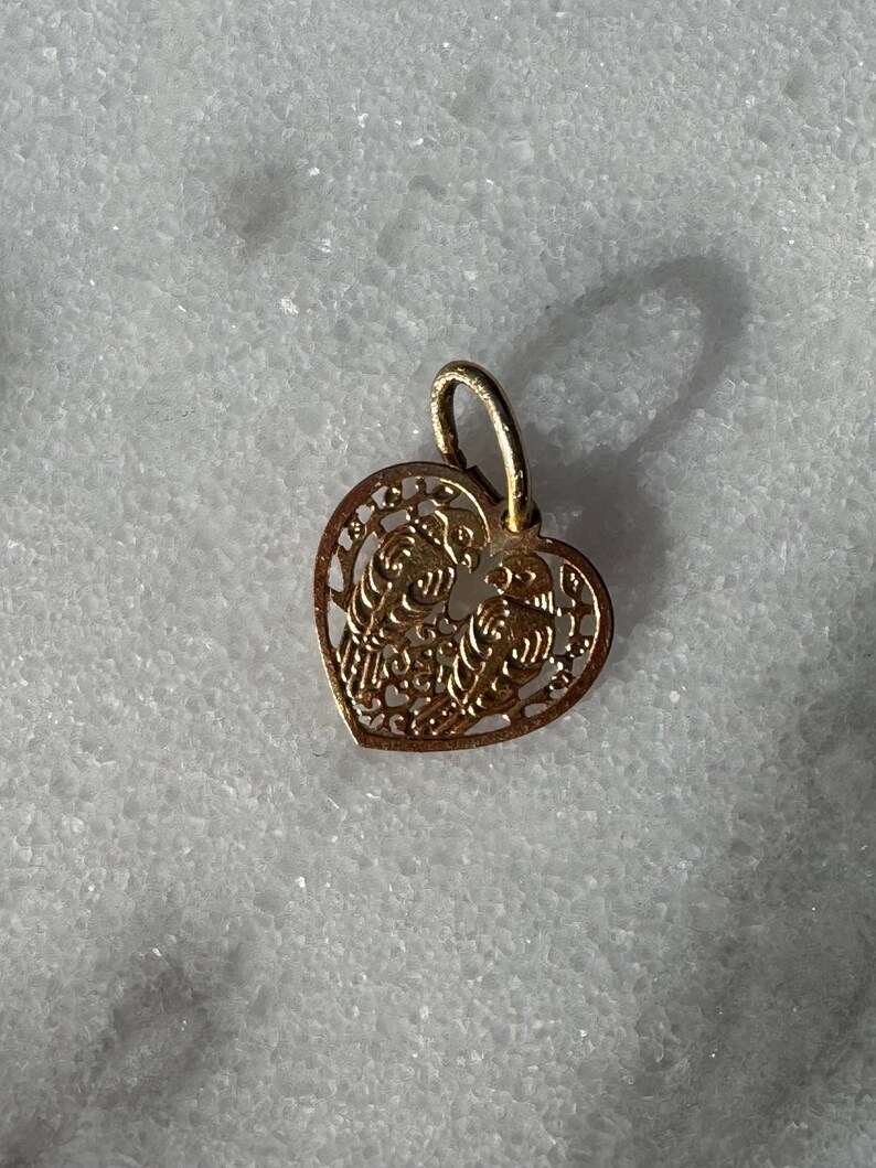 Puede incluir: Un colgante de coraz&oacute;n de oro con un delicado dise&ntilde;o de filigrana. El colgante presenta dos figuras entrelazadas dentro del coraz&oacute;n.