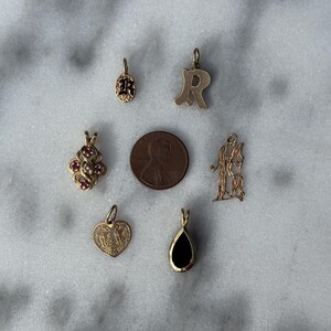 Puede incluir: Una colecci&oacute;n de siete dijes de oro, que incluyen un coraz&oacute;n, una l&aacute;grima, una letra R, un tr&eacute;bol con piedras rojas, un monograma y dos dijes redondos con esmalte negro. Los dijes est&aacute;n dispuestos sobre una superficie de m&aacute;rmol blanco.