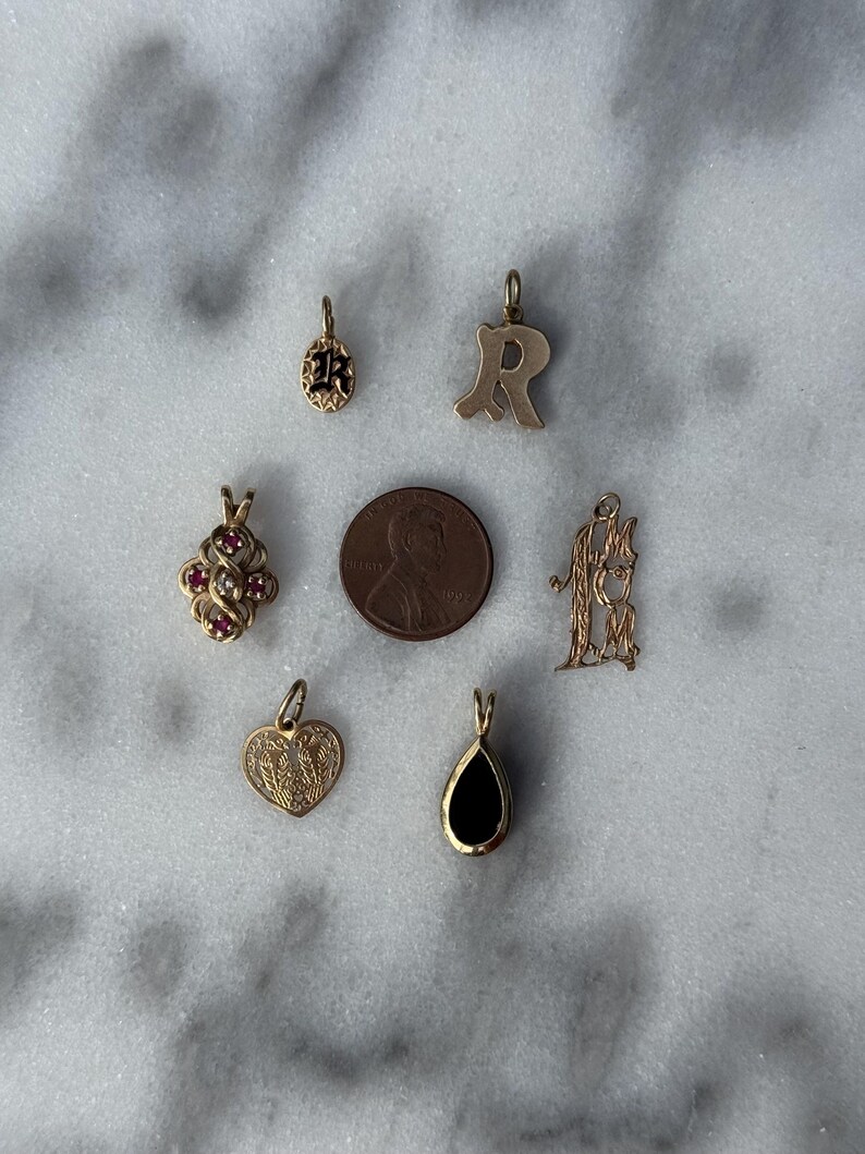 Puede incluir: Una colecci&oacute;n de siete dijes de oro, que incluyen un coraz&oacute;n, una l&aacute;grima, una letra R, un tr&eacute;bol con piedras rojas, un monograma y dos dijes redondos con esmalte negro. Los dijes est&aacute;n dispuestos sobre una superficie de m&aacute;rmol blanco.