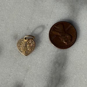 Puede incluir: Un amuleto de coraz&oacute;n de oro con un dise&ntilde;o intrincado. El amuleto es del tama&ntilde;o de una moneda de un centavo de d&oacute;lar estadounidense.