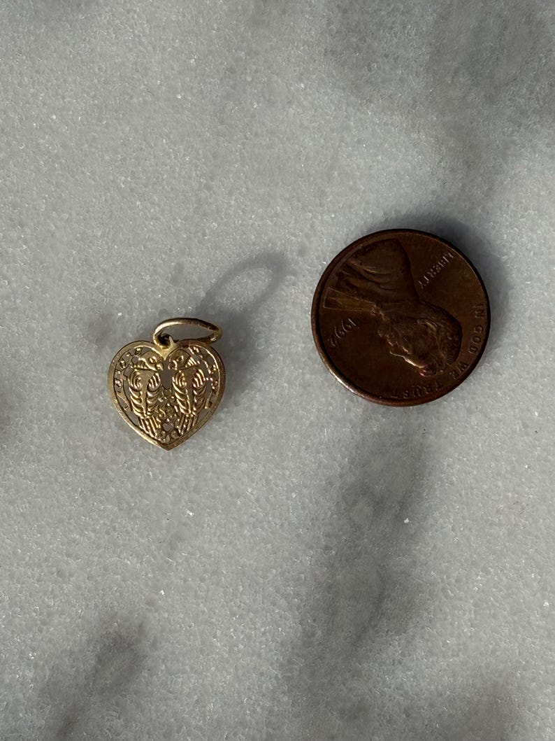 Puede incluir: Un amuleto de coraz&oacute;n de oro con un dise&ntilde;o intrincado. El amuleto es del tama&ntilde;o de una moneda de un centavo de d&oacute;lar estadounidense.