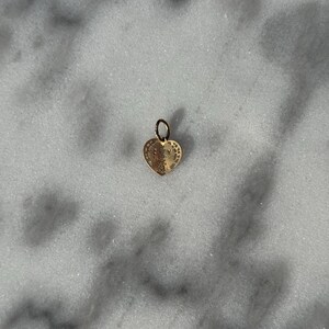 Puede incluir: Un charm de coraz&oacute;n de oro con detalles intrincados. El charm tiene un bucle en la parte superior para sujetarlo a un collar o pulsera.