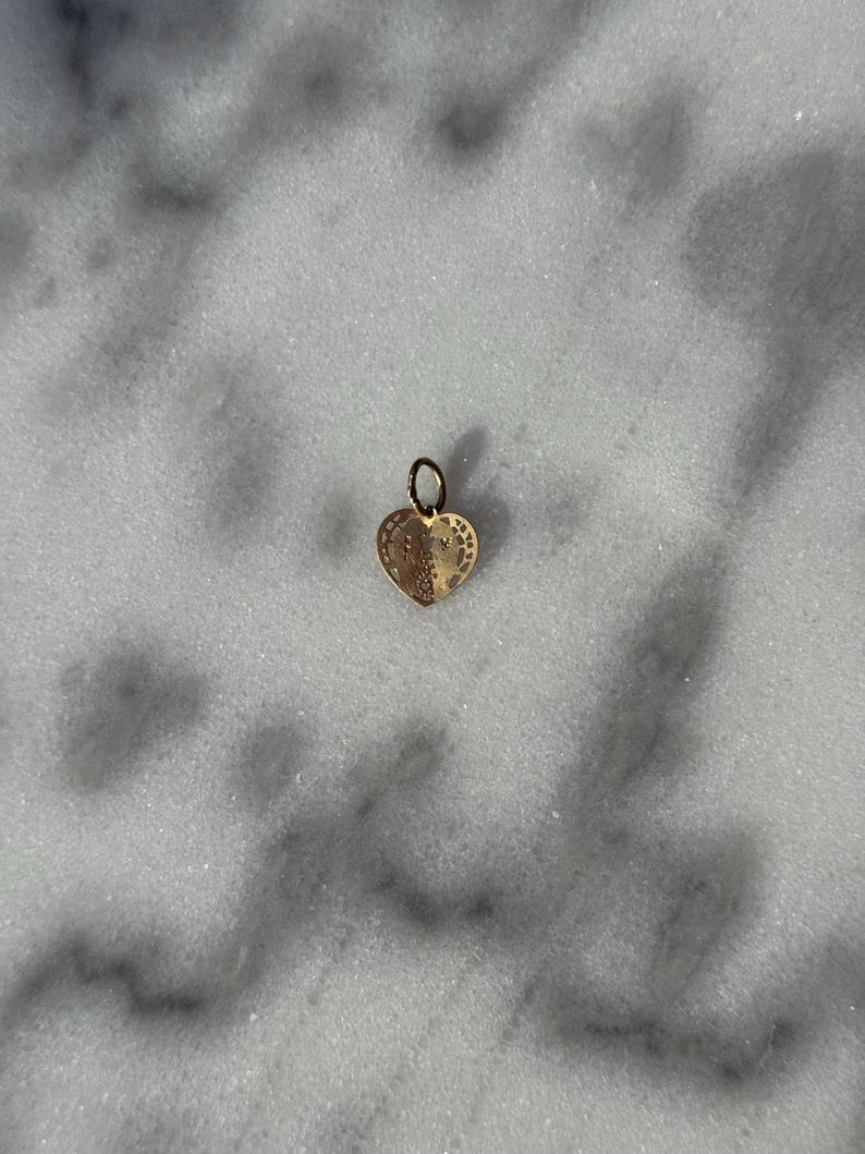 Puede incluir: Un charm de coraz&oacute;n de oro con detalles intrincados. El charm tiene un bucle en la parte superior para sujetarlo a un collar o pulsera.