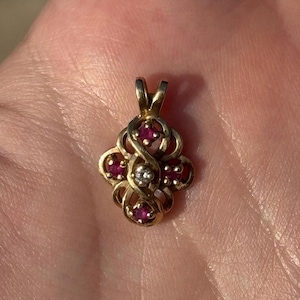 Puede incluir: Un colgante de oro con un diseño delicado que presenta cinco piedras preciosas rojas y una sola piedra blanca en el centro.
