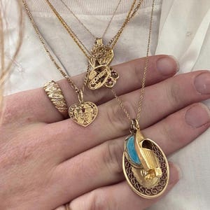 Puede incluir: Primer plano de una mano que lleva varias cadenas de oro con diferentes colgantes. Los colgantes incluyen un coraz&oacute;n, una peque&ntilde;a figura y un colgante m&aacute;s grande con un dise&ntilde;o azul y blanco. La persona tambi&eacute;n lleva dos anillos de oro, uno con una piedra verde y otro con un dise&ntilde;o retorcido.