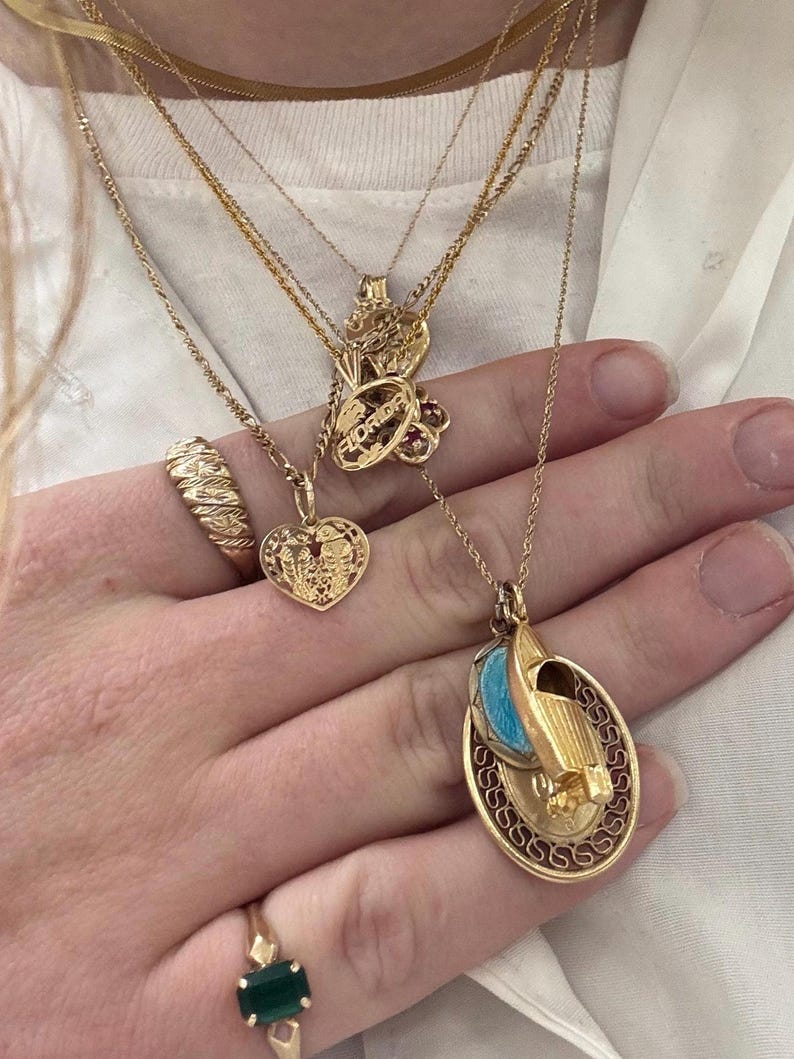 Puede incluir: Primer plano de una mano que lleva varias cadenas de oro con diferentes colgantes. Los colgantes incluyen un coraz&oacute;n, una peque&ntilde;a figura y un colgante m&aacute;s grande con un dise&ntilde;o azul y blanco. La persona tambi&eacute;n lleva dos anillos de oro, uno con una piedra verde y otro con un dise&ntilde;o retorcido.