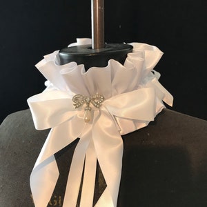 Peut inclure: Un col blanc en satin froissé avec un grand nœud et de longs rubans. Une petite broche scintillante avec une perle pendante accentue le nœud. Le col est présenté sur un mannequin noir.