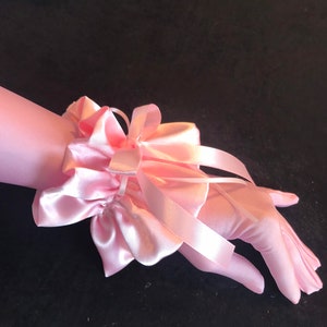Peut inclure: Une paire de gants en satin rose clair avec un détail de poignet froncé assorti et un grand nœud. Les gants sont présentés sur une surface sombre, mettant en valeur la texture et la couleur délicates.