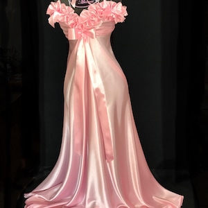 May include: A pink satin nightgown with a ruffled neckline and a bow at the waist. The gown has a long, flowing skirt and is trimmed with lace.