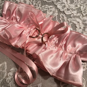 May include: A light pink satin wedding garter with ruffled edges and gold-colored ring accents. The garter features attached ribbons and is designed for a bridal occasion. The background is a gray lace fabric.