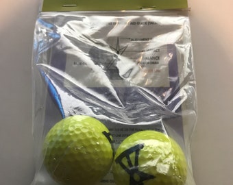 May include: Two yellow golf balls with black markings. One ball has a black target design and the other has a black line and a black circle. The balls are in a clear plastic bag with a green and white label that says "PLAY NOW FOCUS TATAG" and "TARGET ALIGNMENT TRAINING AID GUIDE (TATAG)".