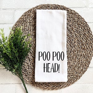 Puede incluir: Toalla de cocina blanca con texto negro que dice "Poo Poo Head!" La toalla está sobre un mantel individual tejido de color marrón.