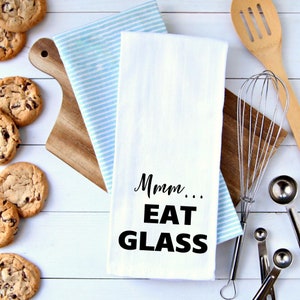 Puede incluir: Toalla de cocina blanca con texto negro que dice "Mmm... EAT GLASS". La toalla est&aacute; doblada y colocada sobre una superficie de madera blanca con una toalla a rayas azules y blancas debajo. Hay galletas con chispas de chocolate y una tabla de cortar de madera en el fondo.