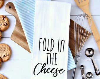 May include: White kitchen towel with black text that reads "Fold in the Cheese". The towel is folded in half and is lying on a white wooden surface. There are kitchen utensils and cookies in the background.