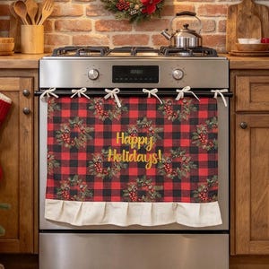 May include: A rectangular oven towel featuring a red and black buffalo plaid pattern with a repeating wreath design and the text Happy Holidays in gold script. The towel has a ruffled cream-coloured bottom edge and is tied to the oven handle.