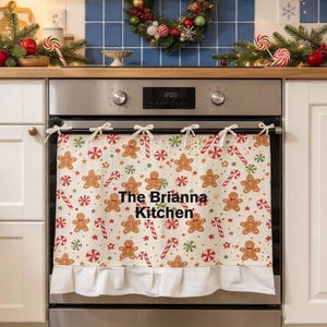 May include: A cream-coloured oven towel featuring a repeating pattern of gingerbread men, red and green candy canes, and peppermint swirls. The towel is tied to the oven handle with white fabric bows and includes the text The Brianna Kitchen.
