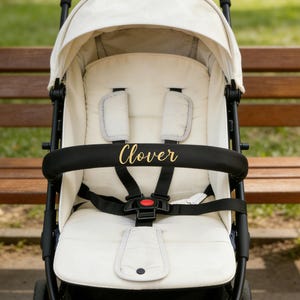 May include: A cream-colored stroller with a black frame and accents. The stroller features a canopy, a padded seat, and a safety harness. The word "Clover" is printed in gold lettering on the black safety bar. The stroller is positioned in front of a wooden bench.