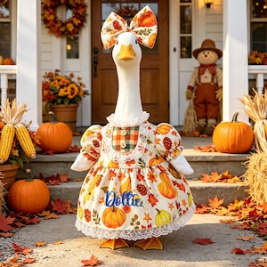 May include: A white goose figurine wearing a cream-colored dress with a fall-themed print of pumpkins, leaves, and pinecones. The dress features a plaid bib, lace trim, and the name Dollie in blue script. A matching bow sits atop the goose's head.