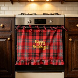 May include: A red and black tartan oven towel featuring the gold embroidered text Happy Holidays! The towel is attached to the oven handle with several fabric ties and includes a ruffled bottom edge, hanging over a stainless steel oven door.