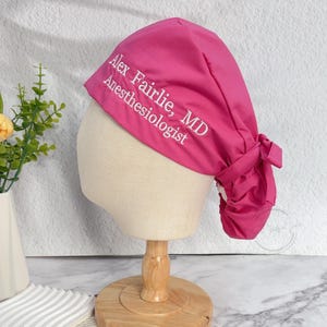 May include: A magenta surgical scrub cap with white embroidered text. The cap is tied at the back and displayed on a mannequin head. The text reads "Alex Fairlie, MD Anesthesiologist".