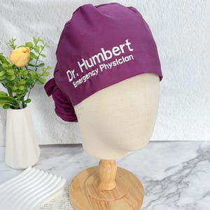 May include: A deep purple surgical scrub cap with the text "Dr. Humbert Emergency Physician" in white. The cap is displayed on a mannequin head with a wooden stand. The background includes a white vase with greenery and a marble-patterned surface.
