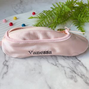 Puede incluir: Estuche de satén rosa claro con forma de zapatilla de ballet, con cierre de cremallera y el nombre "Vanessa" bordado en negro. Tiene una cinta y un tirador de cremallera dorado.