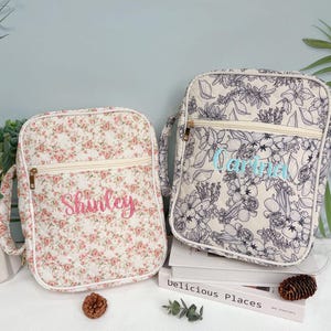 May include: Two quilted fabric bags. One bag features a floral pattern with pink flowers and the name "Shirley" embroidered in pink. The other bag has a black and white floral design with the name "Carina" embroidered in blue.