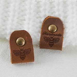 May include: Two brown leather cord ends with a brass button and a laser engraved bee design. One cord end has the word "Handmade" engraved above the bee.