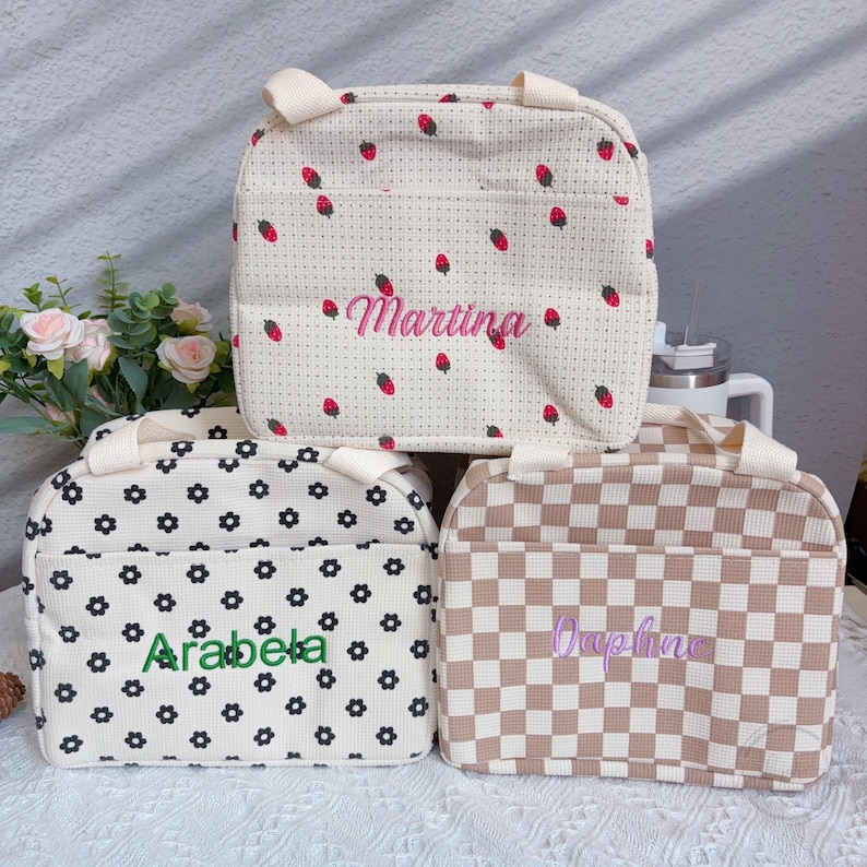 May include: Three personalized lunch bags. One is cream-colored with red strawberries and the name "Martina" embroidered in pink. Another is white with black flower patterns and the name "Arabela" in green. The third is tan and white checkered with the name "Daphne" in purple.
