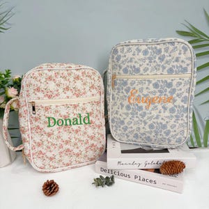 Puede incluir: Dos bolsas con estampado floral y nombres bordados. Una bolsa presenta un estampado floral rosa y rojo con el nombre "Donald" en verde. La otra bolsa tiene un estampado floral azul con el nombre "Eugene" en naranja. Ambas bolsas tienen cierre de cremallera.