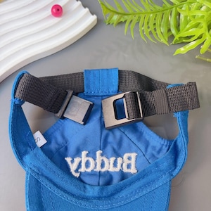 Peut inclure: Casquette de baseball bleue pour animaux de compagnie, avec le mot "Buggy" brod&eacute; en blanc. Elle est dot&eacute;e d'une sangle noire avec une boucle et d'un d&eacute;tail de sangle bleu. La casquette est con&ccedil;ue pour les petits animaux.