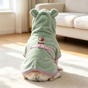 May include: A sage green hooded pet bathrobe with bear ears on the hood. The back features an embroidered red heart with a paw print and the name Bella in red text. A light pink adjustable strap with a plastic buckle secures the robe around the waist.