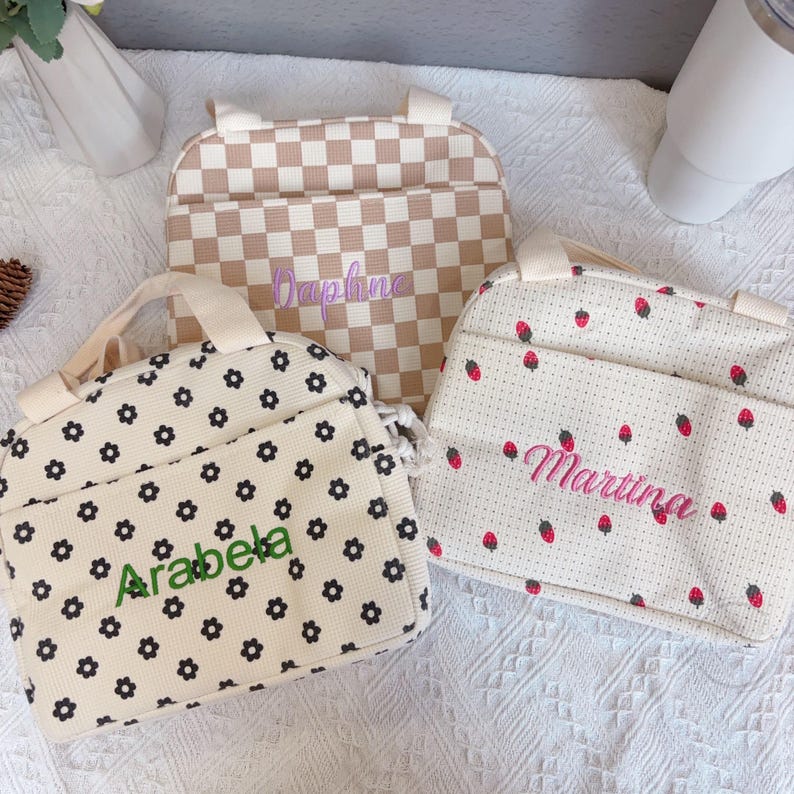 May include: Three personalized tote bags. One bag has a beige and white checkered pattern with the name "Daphne" embroidered in purple. Another has a floral pattern with the name "Arabela" in green. The third bag has a strawberry pattern with the name "Martina" in pink.