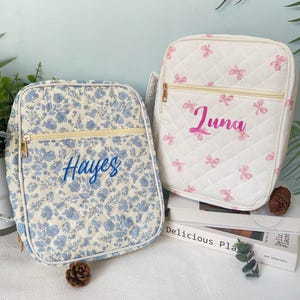 May include: Two quilted, personalized bags. One features a blue floral pattern with "Hayes" embroidered in blue. The other is white with pink bow accents and "Luna" in pink script. Both have gold zippers and are rectangular.