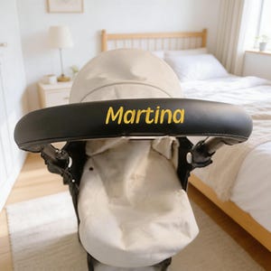 Könnte beinhalten: Ein Kinderwagen mit einem schwarzen Griff, auf dem der Name "Martina" in goldfarbenen Buchstaben steht. Der Kinderwagen hat ein beiges Verdeck und einen passenden Sitz. Das Bild ist in Innenräumen aufgenommen, mit einem Bett und einer Lampe im Hintergrund.