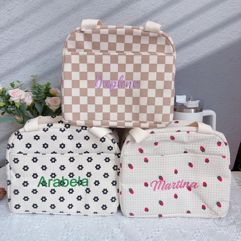 May include: Three lunch bags with different patterns and names. The top bag has a brown and white checkered pattern with the name "Daphne" embroidered in purple. The middle bag has a white background with black flower patterns and the name "Arabela" in green. The bottom bag has a white background with red strawberry patterns and the name "Martina" in pink.