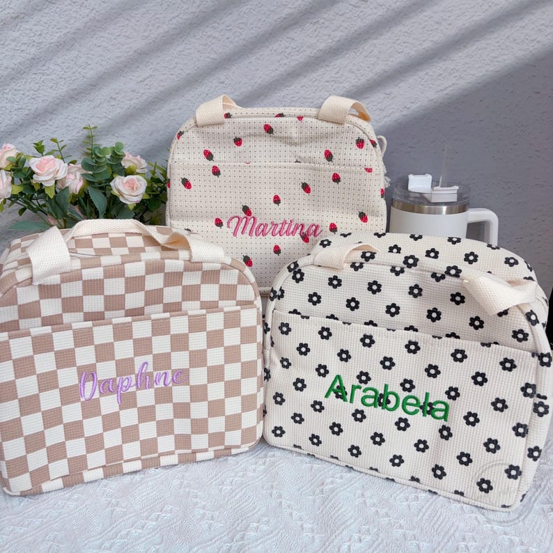 May include: Three personalized lunch bags with different patterns. One bag has a beige and white checkerboard pattern with the name "Daphne" embroidered in purple. Another has a strawberry pattern with the name "Martina" in pink. The third bag has a floral pattern with the name "Arabela" in green.