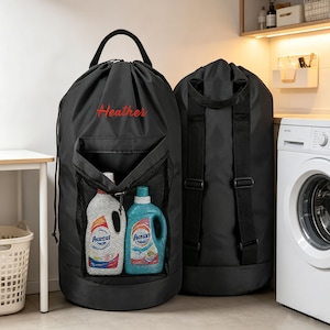 May include: Two black cylindrical laundry bags with drawstring closures and top handles. The front bag features the name Heather in red script and a mesh pocket containing two plastic detergent bottles. The back bag has adjustable shoulder straps.