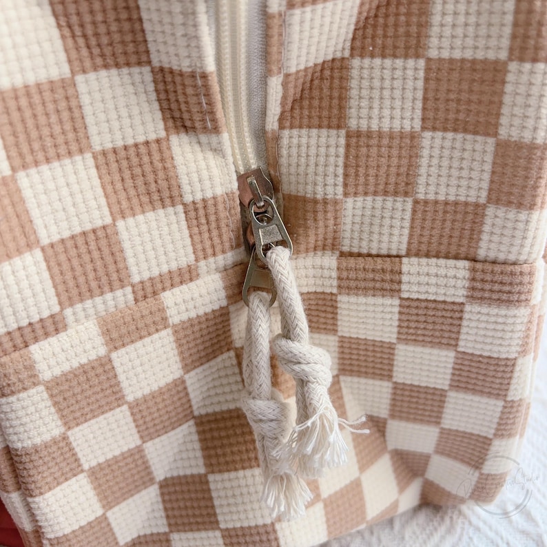 May include: Close-up of a beige and white checkered fabric with a silver zipper. The zipper pull is a white rope with tassels. The fabric has a waffle-like texture.